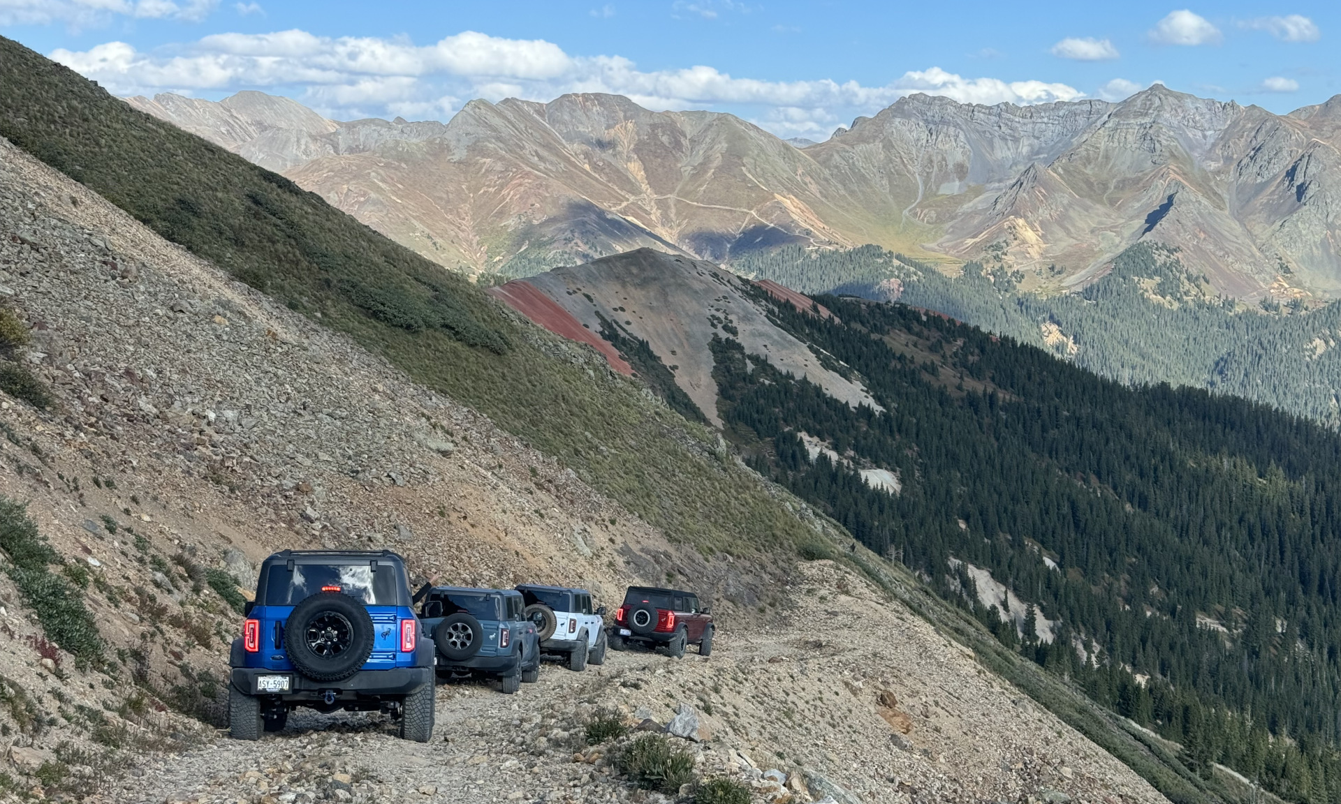 A convoy of off-road vehicles traveling on a mountain trail with spectacular mountain peaks and valleys in the background, representing endless adventure possibilities.