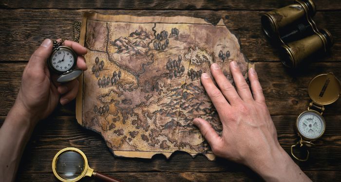 Hands examining an aged treasure map on a wooden surface with vintage navigation tools including compass, binoculars, and pocket watch, representing the authentic treasure hunting experience.